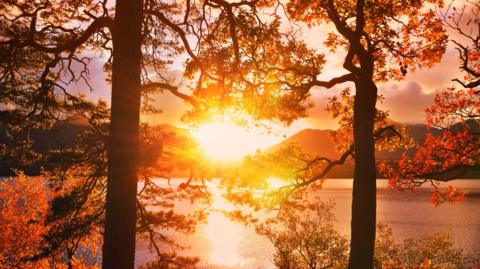 The Sun sets behind hills and casts a golden glow over a lake, with trees silhouetted in the foreground