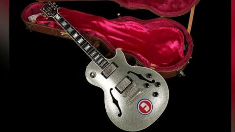 The silver sparkly guitar is laid down next to an opened brown guitar case which is lined with red fabric. 