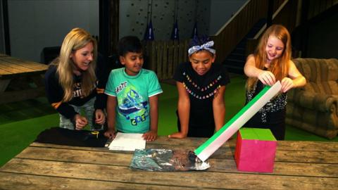 Presenter Naomi Wilkinson and three children. The children are rolling a marble down a sheet of rolled card onto a wooden table.