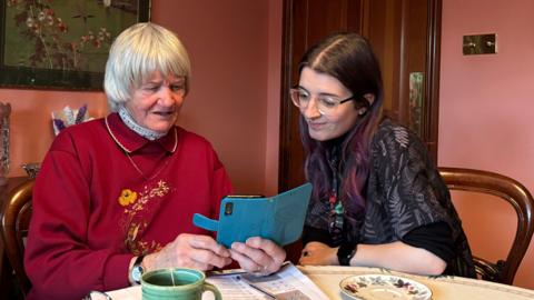 Two women are sitting at a table and looking at a smartphone. The phone is in a blue case. The woman on the left is elderly, with short white hair. She is wearing an embroidered red jumper. The woman on the right is younger. She is wearing a dark shirt and has clear plastic glasses on, with long dark hair that is tinted pink at the ends.