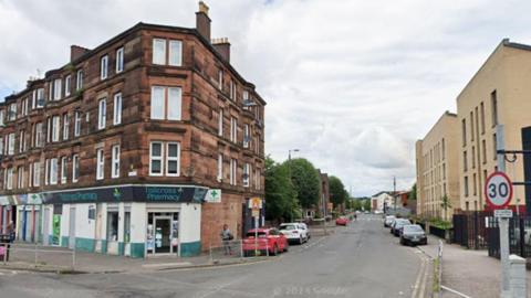 A junction on a city street, with rows of redbrick flats down one side of the street, above a pharmacy