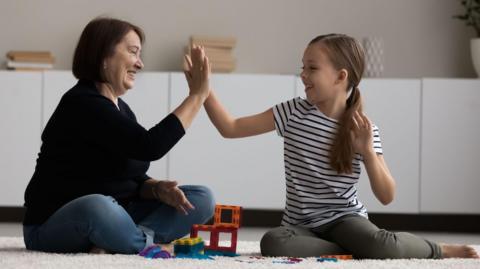 A stock image of a woman high-fiving a smiling young girl as they play with toys together on a rug.