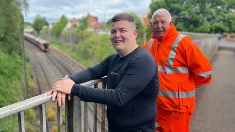 Two men stand on a railway bridge smiling at the camera. The man on the right wears an orange hi-vis National Rail jacket while the man on the left wears a black long-sleeved shirt and jeans. Below on the railway track, a train is on the line.