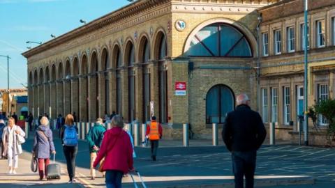 The exterior of Cambridge Station with a number of people walking outside. It is a yellow-brick building with large glass-panelled arches. 