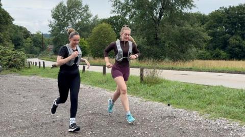 Image shows two females running. One is dressed in all black. She has a black short-sleeved top and black leggings on with a grey running vest over the top. She has black Nike trainers on. Her hair is in a bun. The second officer has turquoise trainers on. Burgundy shorts and a brown long-sleeved tight fitting top. She has a grey running vest over the top. Her hair is in a ponytail. They are in a carpark with four cars parked alongside them. Behind them are a number of trees, grass and a main road. 
