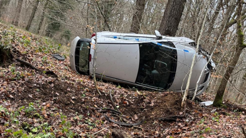 A damaged silver car lying on its side in woodland with trees surrounding it