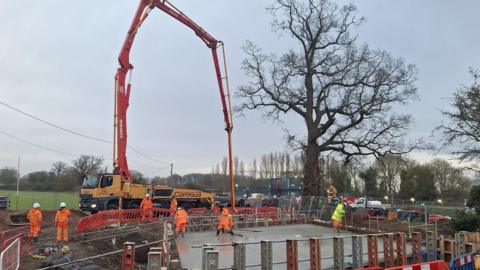 Workers and machinery at the site of the construction of the new bridge. The area has been cordoned off and mud surrounds the site.