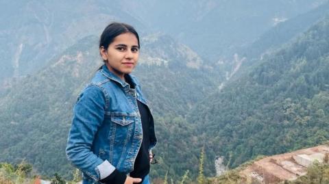 Harshita Brella, who has tied-back black hair and is wearing a denim jacket and a black top. She is posing for a photo in a high spot with tree-covered mountains behind her. 