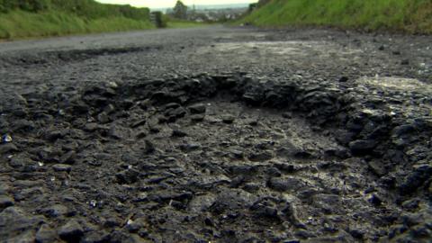 A hole in a dark gravel road up close with a road continuing in the background surrounded by fields