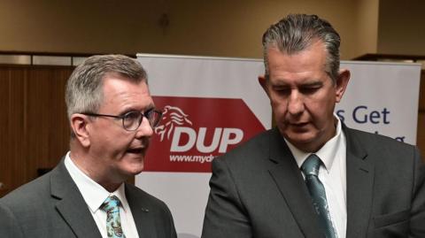 Jeffrey Donaldson and Edwin Poots pictured side by side. Both wearing grey suits. DUP poster in background.