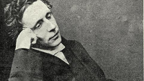 Archive photo of Lewis Carroll seated in a relaxed pose, wearing formal Victorian-era clothing including a dark suit and white shirt. He is resting one arm on the chair and the other near the face. 
