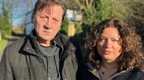 A joint headshot image of David and Karen Yates, standing together in a sunny, rural setting