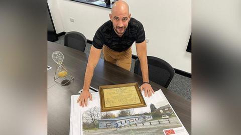 Dale Gaucas, wearing a black top and beige trousers, is leaning over a large photograph of the boxing club built in his old Darnhill estate.   