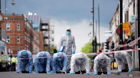 Six police forensic officers on their hands and knees in a line on Kingsland High Street, , near to the scene of the shooting. In front of them is red and white police tape, and a seventh forensic officer stands behind the group.