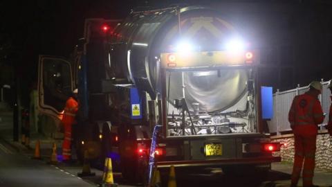 Tanker parked on side of road with workers in orange overalls standing beside it - the tanker has its lights on and it is night.