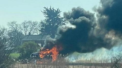 A large cloud of black smoke rising from a tractor which is on fire. Grass and a building can be seen in the background.