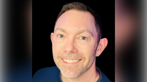 A man with short black hair and a blue top smiles as he stands in front of a black background.