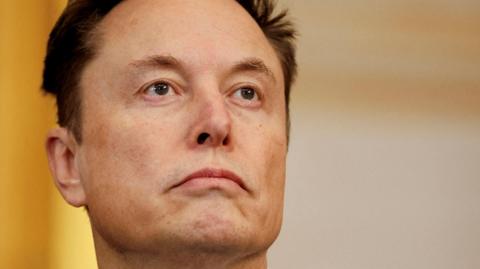 Elon Musk, standing against a light yellow background, looking at something beyond the camera