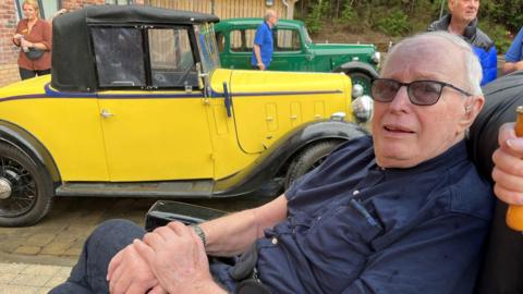 An elderly man with short white hair, glasses, a blue shirt, with his hands clasped, sits in a wheelchair and looks at the camera. Behind him is an old yellow car with a black roof. 
