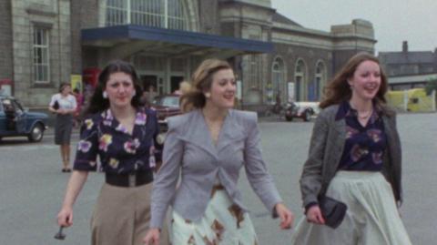 Three young women in 70s fashion, walking out of Margate train station.