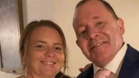 A woman smiling next to a man, who is wearing a white shift, suit and pink tie