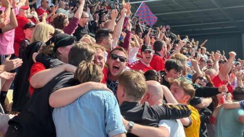 A crowd of happy football fans embrace and cheer, in a football stadium.
