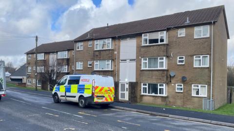 A police van with 'heddlu' written on the side is parked outside a block of flats on High Street, Hirwaun.