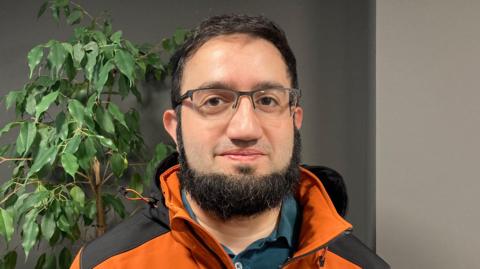 Imraan Moolla smiling at the camera. He is wearing glasses and has a black and orange coat on.