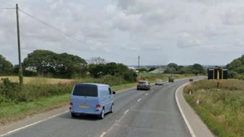 The image shows a road with two lanes and a few vehicles driving away from the camera. The vehicle closest to the camera in a light blue van.