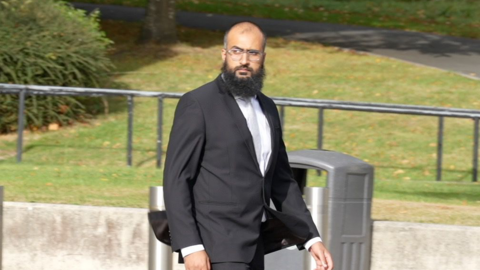A bald man with a beard, in a black suit and white shirt walking. 