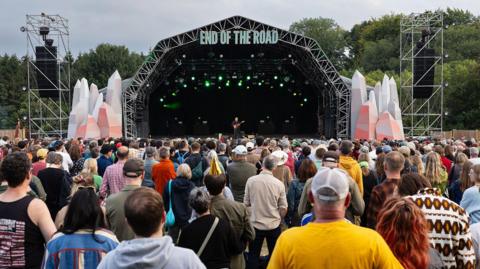 A festival stage with lettering saying 'End Of The Road' on top, with a large crowd of people watching with their backs to the camera. A man stands on stage holding a red guitar. It's a cloudy day and there are trees behind the stage.