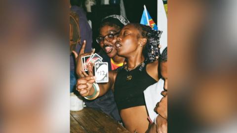 Two young women playing uno, one of them is showing a card with '+12' written on it