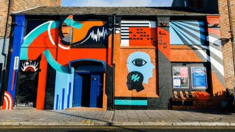 A brick building on a street is painted with colourful murals of shapes and outlines of people's heads. 