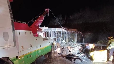 A fire-damaged bus on the side of a road. There is a lorry in front of it using a crane to move it. There are two people on the right