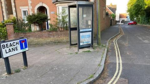 Road sign on left of pavement along with phone box and house behind a lane leads into the distance with a red car parked towards the end.