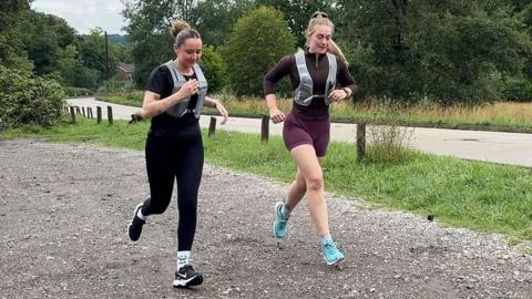 Image shows two females running. One is dressed in all black. She has a black short-sleeved top and black leggings on with a grey running vest over the top. She has black Nike trainers on. Her hair is in a bun. The second officer has turquoise trainers on. Burgundy shorts and a brown long-sleeved tight fitting top. She has a grey running vest over the top. Her hair is in a ponytail. They are in a carpark with four cars parked alongside them. Behind them are a number of trees, grass and a main road.