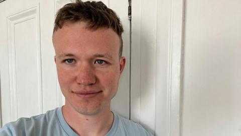 Éanna Mac Cana is smiling at the camera while standing in front of a white door and white wall. He has short, light brown hair and is wearing a light blue t-shirt.