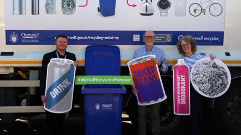 Ian Upstone from Cherwell District Council, councillor Ian Middleton and Julie Meeks of Alupro stand next to a blue recycling bin holding large cutout figures of metal products