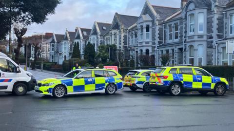 Police cars are seen blocking a road in Plymouth. There are a row of terrace houses behind them.