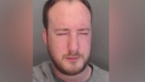 Mugshot of Henderson. He is 30 with light brown hair, a brown beard and is wearing a grey jumper. His eyes are closed.