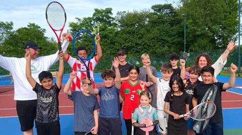 Photo of 15 people smiling and cheering on bright coloured tennis courts. 