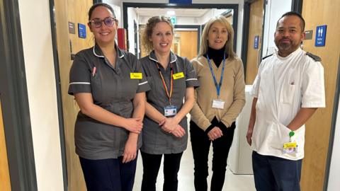 Four hospital workers standing in a hallway in Bath hospital all smiling. Two are wearing grey nurses' outfits, one man is wearing a white uniform with blue trousers and another woman is wearing black trousers and a cream top.