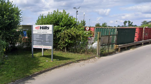 A view of a road on a sunny day with large metal containers for recycling behind a fence, trees on the opposite side. A large sign for Hills waste to the left.