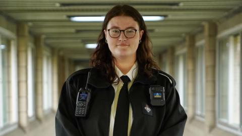 Hollie Peabody at work. They wear a black prison officer's uniform including a black tie and jacket over a white shirt. They have a pager and walkie-talkie attached to their jacket and wear dark-rimmed rectangle glasses. Hollie is pictured in a brutalist corridor with windows on either side and looks straight at the camera. 