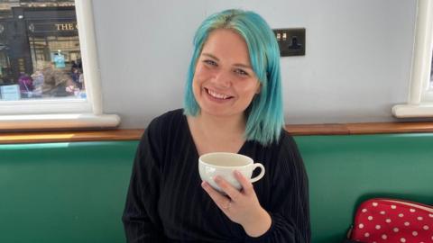 A woman with blue shoulder length hair smiling as she holds up a white mug. She is sitting on a green bench and a shop can be seen behind her. She is wearing a black top.