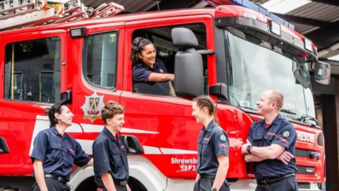 Male and female firefighters stand by a fire engine with one woman in the driver's seat. They are smiling and looking at one another while wearing their blue uniforms.