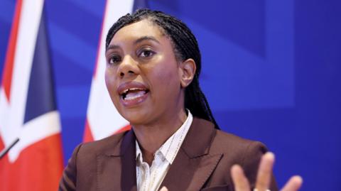 Kemi Badenoch speaking in front of British flag at an event. She is wearing a brown coloured jacket and a white shirt.