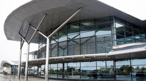 A modern steel and glass building on a rainy day. There are huge glass windows reaching up several storeys high. Cars are reflected in the building.