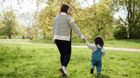 A mum walking and hold her child's hand near trees on a sunny day.