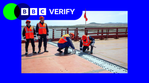 Four workmen wearing bright orange hats and reflective high-vis jackets stand at the centre of a new road bridge between Russia and North Korea. The centrepoint where the two ends of the bridge join is marked by a line of steel plates. Two of the workmen are crouched over the plates, examining them. 